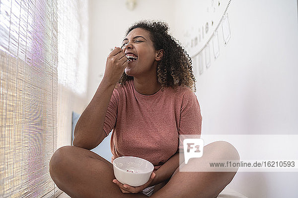 Fröhliche Frau isst Sahne und Erdbeeren  während sie zu Hause an der Wand sitzt