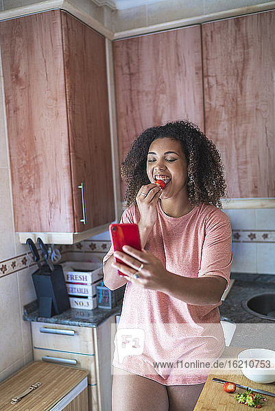 Fröhliche junge Frau  die eine Erdbeere isst  während sie in der Küche einen Videogespräch über ein Smartphone führt