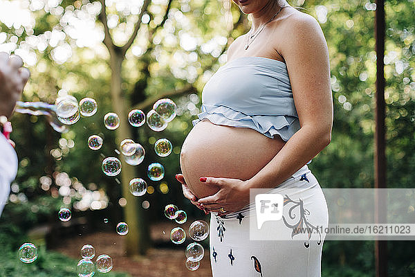Schwangere Frau mit Händen auf dem Bauch steht bei Blasen im öffentlichen Park