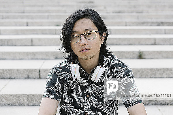 Nahaufnahme eines jungen Mannes mit Brille und Kopfhörern  der auf einer Treppe in der Stadt sitzt