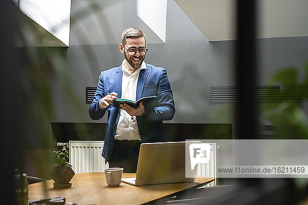 Smiling male professional holding diary while looking at laptop on desk in illuminated coworking space