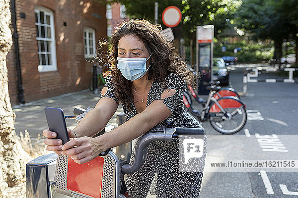 Frau mit Maske  die ein Selfie mit ihrem Smartphone macht  während sie auf einem Fahrrad in der Stadt sitzt