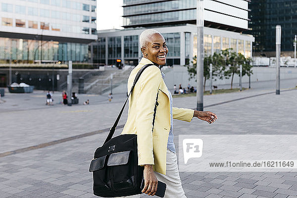 Happy businesswoman with phone and bag walking in city