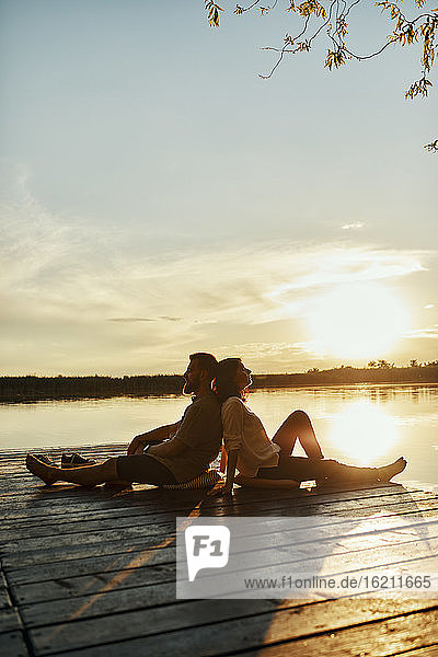 Paar sitzt Rücken an Rücken auf einem Steg an einem See bei Sonnenuntergang
