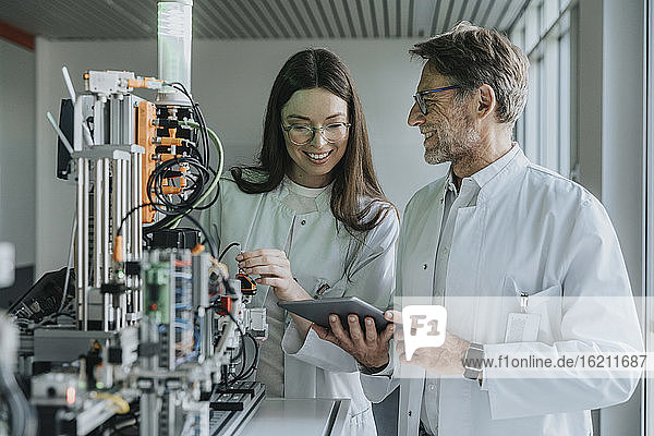 Älterer Mann zeigt einer Kollegin ein digitales Tablet  während er an einer Maschine im Labor steht