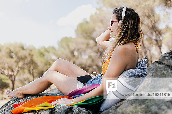Junge Frau mit Regenbogenflagge hört Musik über Kopfhörer  während sie auf einem Felsen sitzt