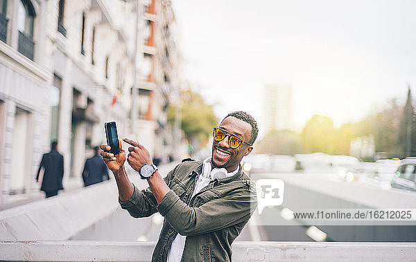 Glücklicher Mann mit Sonnenbrille  der sein Smartphone zeigt  während er in der Stadt steht