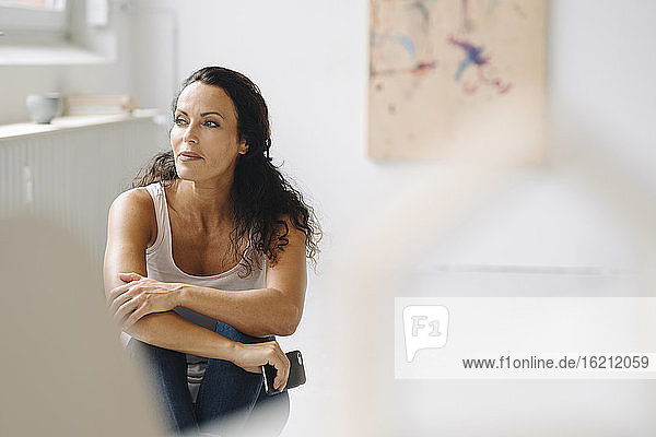 Thoughtful mid adult woman looking away while sitting on floor in loft apartment