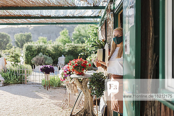 Weiblicher Besitzer mit Maske  der wegschaut  während er am Eingang einer Gärtnerei steht