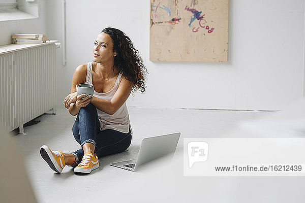 Thoughtful woman holding coffee mug while sitting with laptop on floor at home