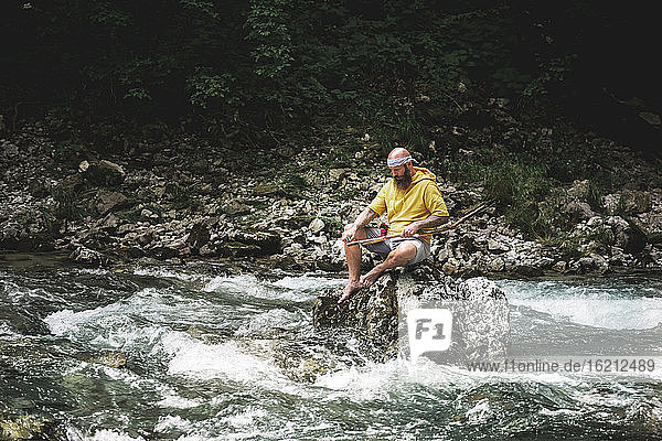 Abenteurer mit Bart sitzt in der Mitte des Flusses auf einem Stein und schnitzt an einem Stück Holz
