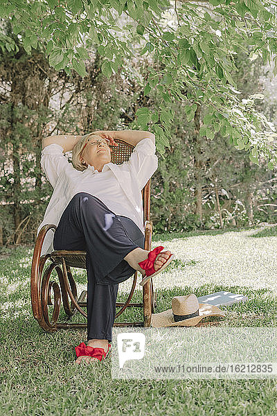 Nachdenkliche ältere Frau mit Händen hinter dem Kopf entspannt auf einem Stuhl im Hof