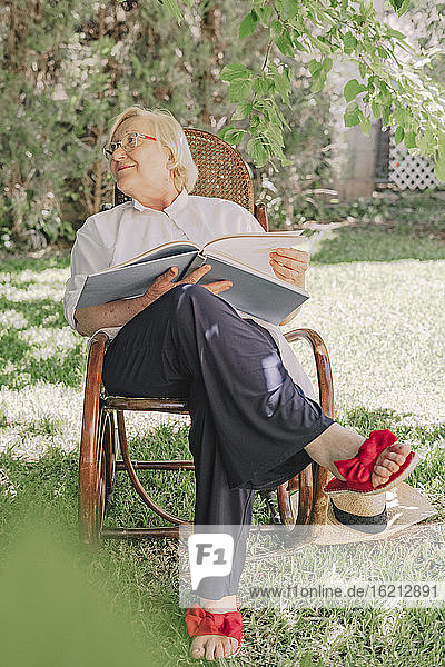 Ältere Frau hält ein Buch und schaut weg  während sie sich auf einem Stuhl im Garten entspannt