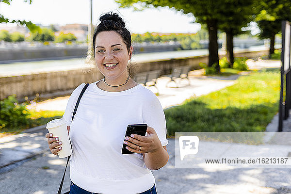 Porträt einer lächelnden  kurvenreichen jungen Frau mit Getränk zum Mitnehmen und Mobiltelefon in der Stadt