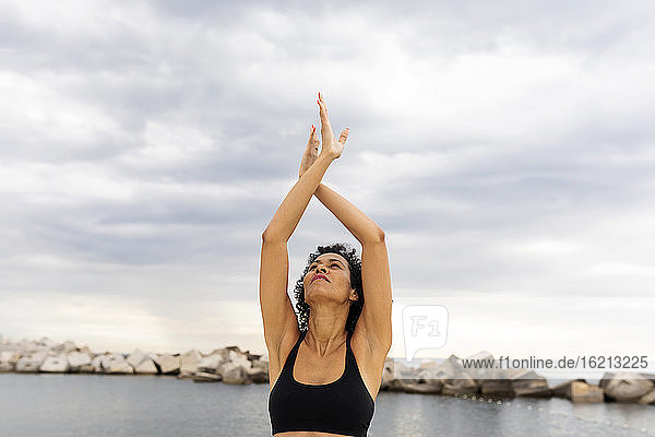 Smiling mid adult woman with arms raised exercising against sea and cloudy sky