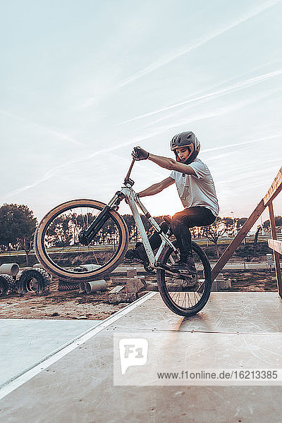 Junger Mann mit Helm fährt Fahrrad auf einer Rampe im Park gegen den Himmel bei Sonnenuntergang