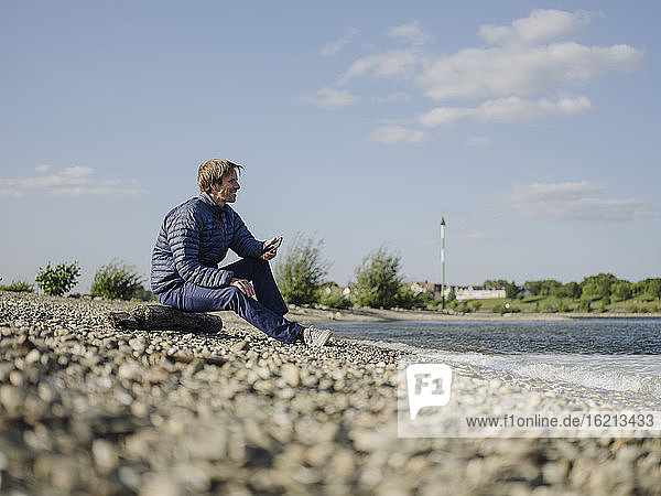 Mature man wearing warm clothing looking at Rhine river while sitting on land against sky