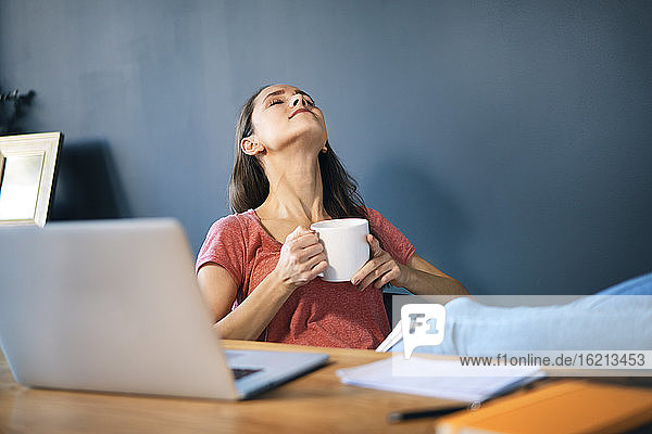 Unternehmerin hält Kaffeetasse  während sie sich am Schreibtisch im Heimbüro entspannt