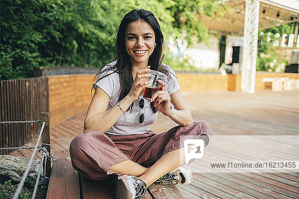 Lächelnde junge Frau  die ein Erfrischungsgetränk in der Hand hält und auf einer Bank im Park sitzt
