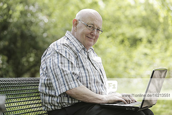 Deutschland  Nordrhein-Westfalen  Köln  Porträt eines älteren Mannes mit Laptop auf einer Parkbank  lächelnd