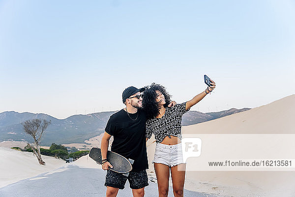 Smiling couple taking selfie while standing on road against clear blue sky