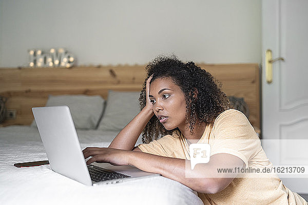 Stressed young woman with head in hand using laptop on bed at home