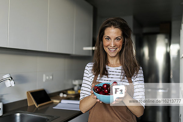 Lächelnde junge Frau  die Kirschen in der Hand hält  während sie in der Küche zu Hause steht