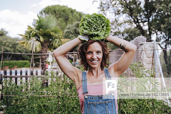 Lächelnde Frau hält Salat auf dem Kopf  während sie im Gemüsegarten steht