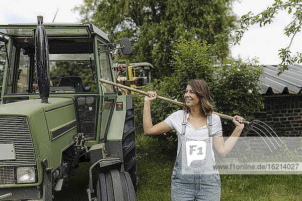 Lächelnde junge Frau mit Heugabel an einem Traktor auf dem Lande