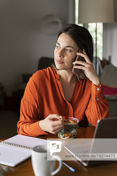 Businesswoman talking on mobile phone while eating salad at home