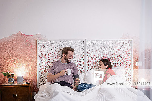 Lächelnder Mann mit Kaffee in der Hand im Gespräch mit seiner Frau  während er sich zu Hause auf dem Bett entspannt