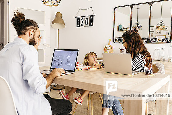 Mann arbeitet am Laptop  während Mutter und Tochter sich im Esszimmer unterhalten