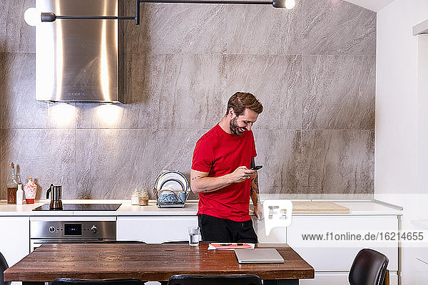 Lächelnder mittelgroßer erwachsener Mann  der ein Smartphone benutzt  während er zu Hause an der Küchentheke steht