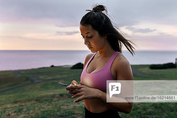 Junge Sportlerin,  die ein Smartphone benutzt,  während sie bei Sonnenuntergang vor dem Himmel steht