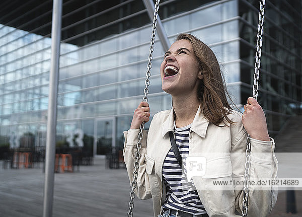 Happy young woman screaming while swinging against modern building in city
