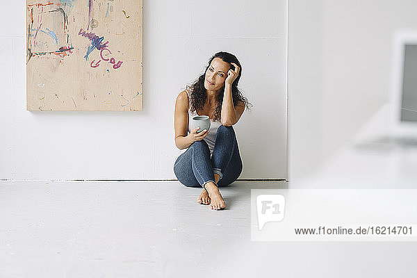 Thoughtful woman holding coffee mug sitting on floor against wall in loft