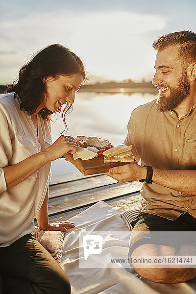 Pärchen beim Picknick auf dem Steg eines Sees bei Sonnenuntergang und beim Essen von Käse