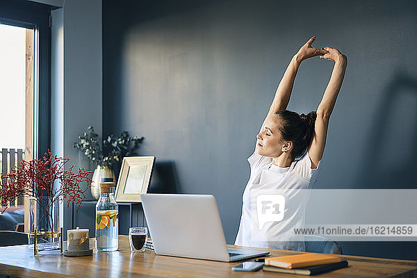 Müde junge Frau  die ihre Arme ausstreckt  während sie im Büro zu Hause am Schreibtisch sitzt