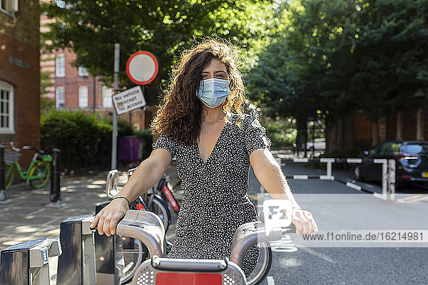 Junge Frau mit Maske sitzt auf einem Fahrrad in einer Parkstation in der Stadt