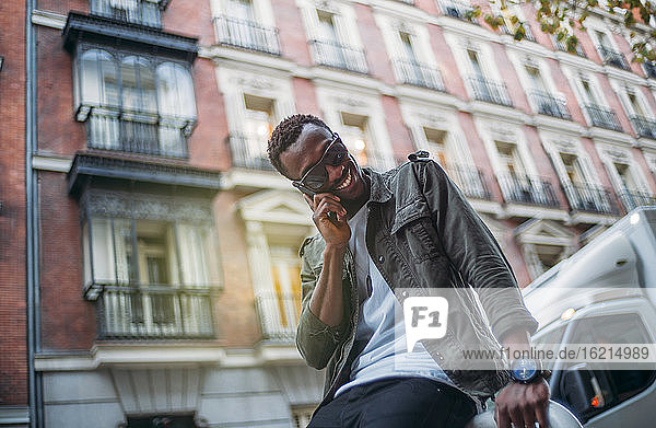 Lächelnder Mann mit Sonnenbrille  der über ein Mobiltelefon spricht  während er an einem Gebäude in der Stadt sitzt