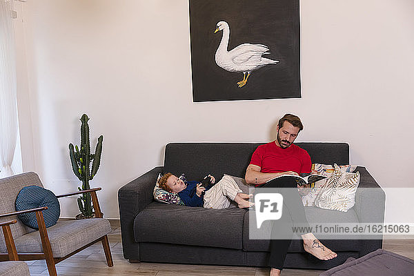 Mann liest eine Zeitschrift  während sein Sohn zu Hause auf dem Sofa mit einem Joystick spielt