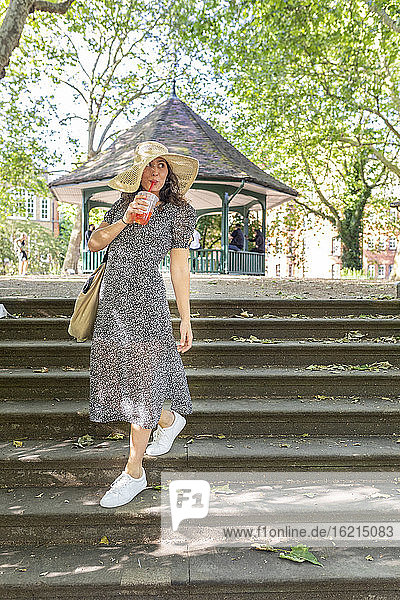 Junge Frau mit Hut  die etwas trinkt  während sie eine Treppe im Park hinuntergeht