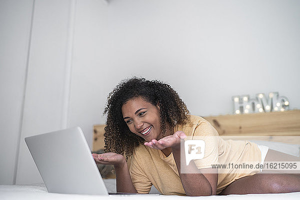Fröhliche junge Frau bei einer Videokonferenz über einen Laptop  während sie zu Hause auf dem Bett liegt