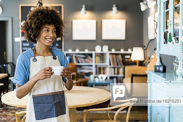 Thoughtful female owner holding coffee cup while standing in modern cafe