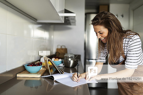 Smiling young woman writing recipe in book while watching video over digital tablet at home