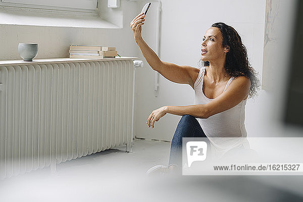 Mittlere erwachsene Frau  die ein Selfie mit ihrem Smartphone macht  während sie zu Hause auf dem Dachboden sitzt
