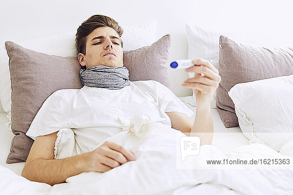 Ein kranker Mann prüft seine Temperatur mit einem Thermometer  während er sich zu Hause auf dem Bett ausruht