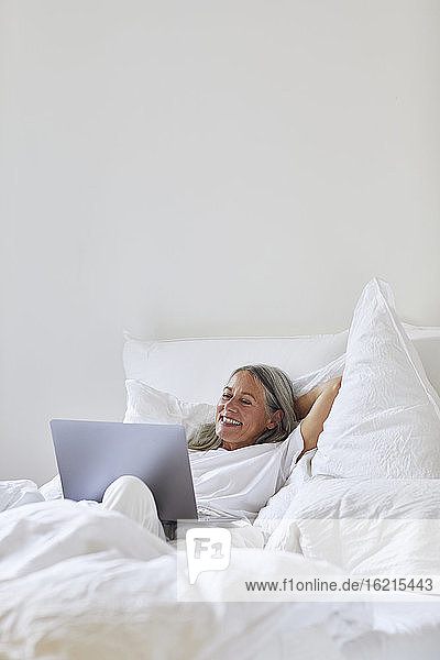 Lächelnde Frau  die einen Laptop benutzt  während sie zu Hause auf dem Bett liegt