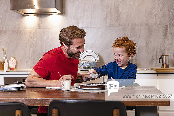 Glücklicher Vater betrachtet seinen süßen Sohn beim Verzieren von Schokoladenkuchen auf dem Esstisch in der Küche