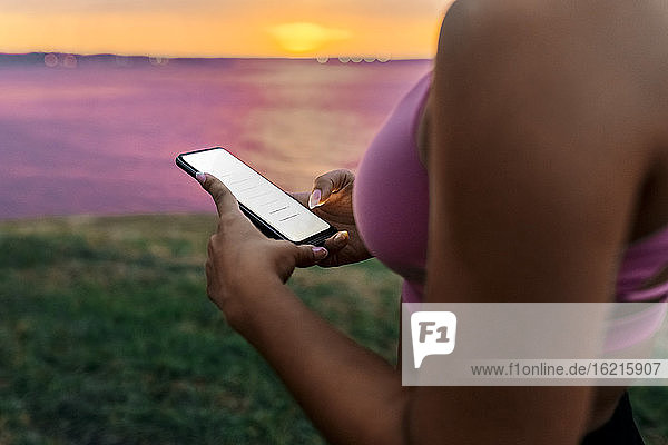 Nahaufnahme einer fitten Frau  die bei Sonnenuntergang ein Smartphone benutzt